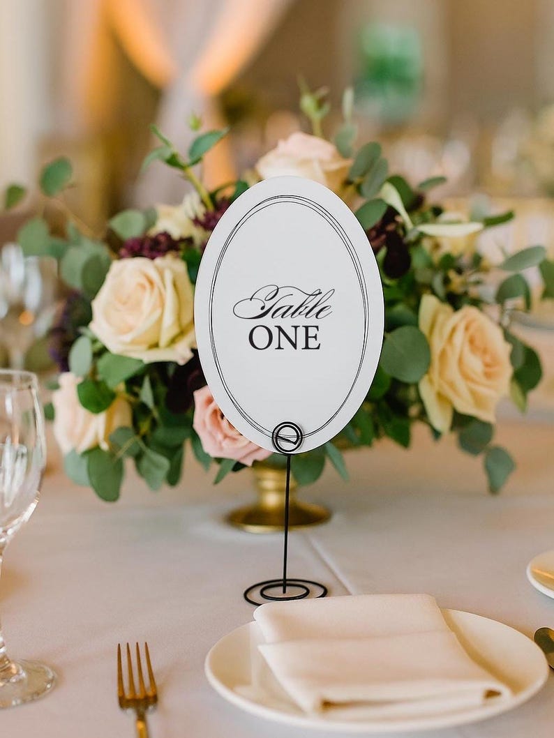 Op de afbeelding: Een bruilofttafelsetting met een bloemstuk en een tafelnummer. Het witte ovale tafelnummer leest "Table ONE" in elegant schrift. Een gevouwen servet en bord staan op de voorgrond.