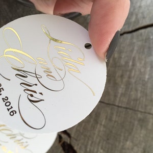 Puede incluir: Etiqueta redonda de papel blanco con letras doradas. La etiqueta dice "Lilla y Chris" y "May 25, 2016".