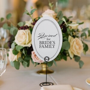 May include: An oval white sign with a thin black border and the text Reserved for BRIDE'S FAMILY in black script and block lettering. The sign is held upright by a thin black wire stand with a circular base, positioned in front of a floral arrangement.