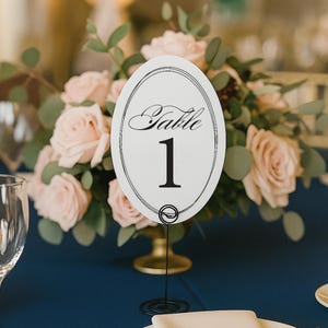 May include: An oval white table number card featuring the word Table in black script above the number 1 in a bold serif font, enclosed by a double-line border. The card is held upright by a thin black wire stand with a coiled circular base.
