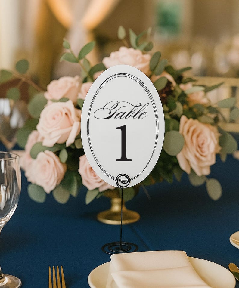 May include: An oval white table number card featuring the word Table in black script above the number 1 in a bold serif font, enclosed by a double-line border. The card is held upright by a thin black wire stand with a coiled circular base.