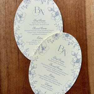 Può includere: Due menu ovali con un design floreale e le iniziali "BA". I menu elencano portate come la zuppa di aragosta spinosa e l'insalata Caesar alla griglia. Il bordo floreale è blu chiaro.