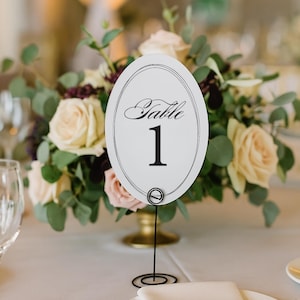 Op de afbeelding: Een witte ovale tafelnummerkaart met het woord "Table" en het nummer "1" in zwart schrift. De kaart wordt vastgehouden door een zwarte draadstandaard en is omgeven door een bloemstuk van crème- en roze rozen en groen. Een gouden vaas houdt de bloemen vast.