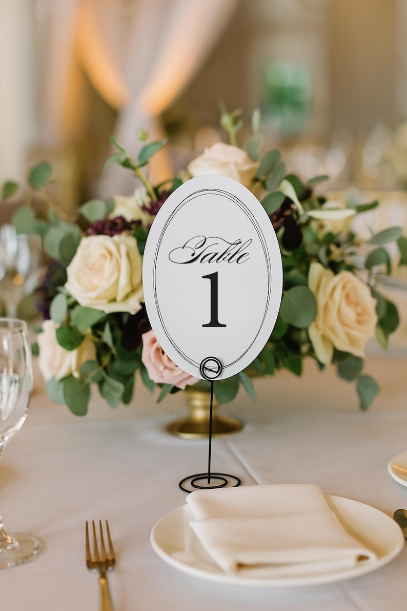 May include: An oval white table number card featuring the text Table 1 in black script and serif font, enclosed by a thin black border. The card is held upright by a thin black metal wire stand with a coiled base, positioned on a table.