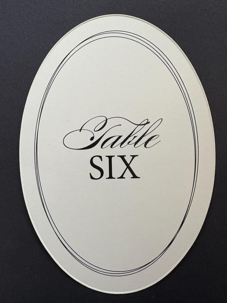 Op de afbeelding: Ovale tafelnummerkaart met het woord "Table" in elegant schrift boven het nummer "SIX" in een vetgedrukte letter. De kaart heeft een witte achtergrond met een dunne zwarte rand.