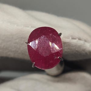 Peut inclure: Une pierre précieuse rouge foncé de taille ovale maintenue dans un sertissage à griffes en métal. La pierre présente une surface à facettes qui reflète la lumière, révélant une teinte cramoisie vibrante et une qualité translucide.