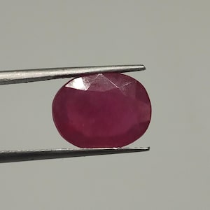 May include: A faceted, oval-shaped ruby gemstone held by tweezers. The ruby is a deep, rich red color, with visible facets that catch the light. The background is a plain, light gray.