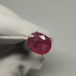 Peut inclure: Une pierre précieuse de rubis facettée, de forme ovale. Le rubis est d'un rouge profond et est maintenu par de petites griffes métalliques. Le fond est de couleur neutre.