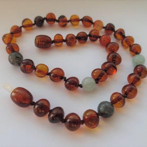 May include: A necklace featuring polished, rounded amber beads in various shades of brown and orange, interspersed with two smooth, pale green beads and two dark grey beads, all strung on a dark cord with a clear plastic screw-style clasp.