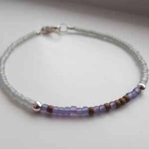 May include: A delicate beaded bracelet featuring a sequence of matte light blue beads, followed by a section of alternating matte purple and brown beads, accented with two polished silver-toned metal beads and a silver-toned lobster claw clasp.