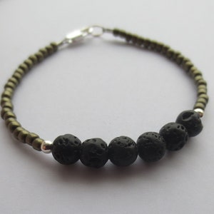 Puede incluir: Una pulsera que presenta una serie de pequeñas cuentas metálicas de color verde oliva y cinco cuentas más grandes de piedra de lava negra porosa. Las cuentas están separadas por pequeños espaciadores de metal plateado y un cierre plateado.