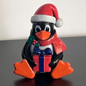 May include: A ceramic penguin figurine wearing a red Santa hat and a red scarf. The penguin has a black body, white chest, and orange beak and feet. It is holding a red gift box with a blue ribbon and bow, accented with green holly leaves.