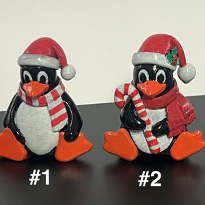 May include: Two small penguin figurines wearing red and white Santa hats. Penguin #1 wears a red and white striped scarf. Penguin #2 wears a solid red scarf and holds a red and white candy cane. Both have orange beaks and feet.