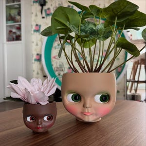 May include: Two doll-face planters, one small and dark-skinned, the other larger with fair skin. The larger planter holds a green plant with large, round leaves. The smaller planter contains a pink flower. Both planters have large, expressive eyes.
