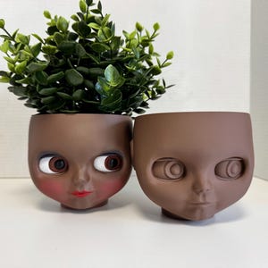 Könnte beinhalten: Zwei braune Blumentöpfe in Form von Puppenköpfen. Einer enthält eine grüne Pflanze. Die Puppenköpfe haben bemalte Merkmale, darunter Augen, rote Lippen und Rouge. Der andere Puppenkopf hat unbemalte Merkmale.