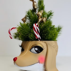 Puede incluir: Una maceta decorativa con forma de perro con temática navideña. La maceta de color canela presenta una nariz roja, mejillas rosadas y sombra de ojos dorada con purpurina. Está llena de pino artificial verde, bastones de caramelo y adornos de reno con purpurina dorada.
