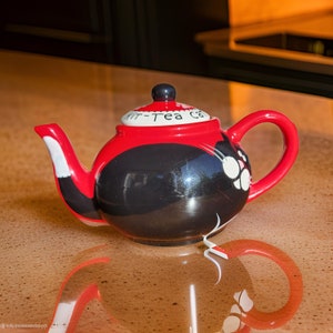 May include: A red and black teapot with a white cat face and the words "Mr-Tea Cat" on the side. The teapot is sitting on a wooden surface.