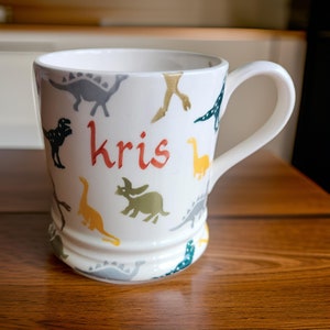 May include: White ceramic mug with a dinosaur pattern and the name "kris" written in orange on the front.