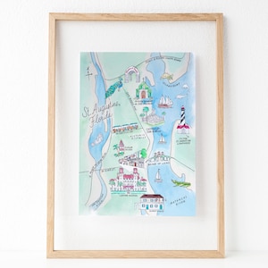 May include: A framed watercolor map of St. Augustine, Florida, featuring hand-drawn illustrations of local landmarks like the Castillo de San Marcos, the St. Augustine Lighthouse, and the Bridge of Lions, all set against a light blue water background.