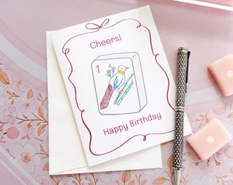 May include: A white greeting card with a maroon border, the text Cheers! and Happy Birthday, and a central illustration of a mahjong tile featuring a bird. A silver textured pen lies next to the card, surrounded by pink mahjong tiles.