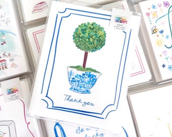 May include: A white greeting card featuring a watercolor illustration of a green topiary tree planted in a blue and white patterned teacup. The card has a blue border and the text "Thank you" printed in blue. A small label reads "EIGHT BLANK CARDS + ENVELOPES".