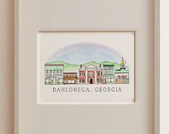 May include: A framed watercolor print featuring a row of buildings in Dahlonega, Georgia, with green hills and a soft blue sky in the background. The text DAHLONEGA, GEORGIA is printed in black capital letters below the illustration.