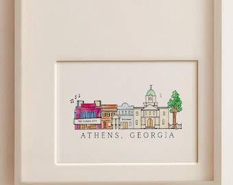 May include: A framed print featuring a colorful hand-drawn illustration of the Athens, Georgia skyline. The artwork depicts a row of buildings, including a clock tower and a green tree, with the text Athens, Georgia printed below.