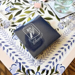 Puede incluir: Una caja azul marino con una ilustraci&oacute;n blanca de un p&aacute;jaro y bamb&uacute;, con la palabra "Cheers!" en cursiva. La caja est&aacute; sobre una superficie con estampado floral con hojas y flores azules y verdes.