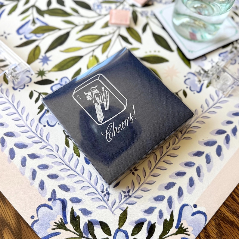 Puede incluir: Una caja azul marino con una ilustraci&oacute;n blanca de un p&aacute;jaro y bamb&uacute;, con la palabra "Cheers!" en cursiva. La caja est&aacute; sobre una superficie con estampado floral con hojas y flores azules y verdes.