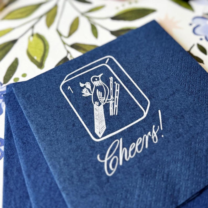 Puede incluir: Servilletas de papel azul marino apiladas, con una ilustraci&oacute;n blanca de un p&aacute;jaro y la palabra "Cheers!" en cursiva. La ilustraci&oacute;n del p&aacute;jaro est&aacute; dentro de un marco blanco.