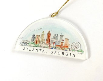 May include: A clear acrylic ornament with a watercolor illustration of the Atlanta, Georgia skyline. The ornament features the city's iconic buildings, including the World of Coca-Cola, the Georgia Aquarium, and the State Capitol. The ornament is decorated with a gold string for hanging.