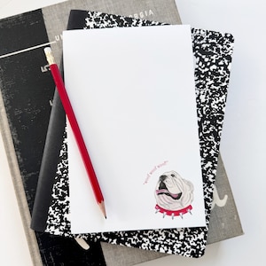 May include: A white notepad with a black and white speckled cover. A red pencil is lying on top of the notepad. The notepad has a small illustration of a white bulldog with a red collar and the text "woof woof" in pink.