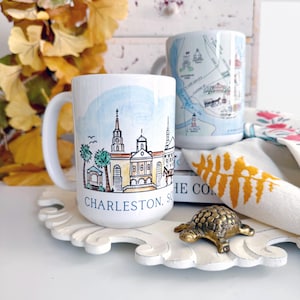 May include: Two white ceramic mugs with illustrations. One mug features a watercolor cityscape of Charleston, South Carolina, with the text "Charleston, SC". The other mug features a map of Charleston, South Carolina. The mugs are on a white tray with a gold turtle figurine.