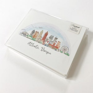 Pode incluir: Um conjunto de oito cart&otilde;es em branco e envelopes com uma ilustra&ccedil;&atilde;o em aquarela do horizonte de Atlanta, Ge&oacute;rgia. A ilustra&ccedil;&atilde;o inclui os ic&ocirc;nicos arranha-c&eacute;us da cidade, uma roda gigante e um arco-&iacute;ris.