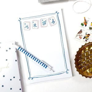 May include: A white notepad with a blue border and Mahjong tile illustrations. A blue and white striped pen rests on the notepad. The notepad is on a white surface with other decorative items.
