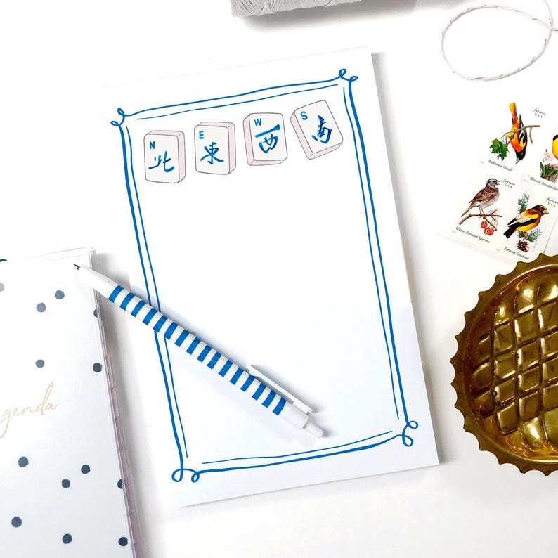 Puede incluir: Un bloc de notas blanco con un borde azul e ilustraciones de fichas de Mahjong. Un bol&iacute;grafo a rayas azules y blancas descansa sobre el bloc de notas. El bloc de notas est&aacute; sobre una superficie blanca con otros elementos decorativos.