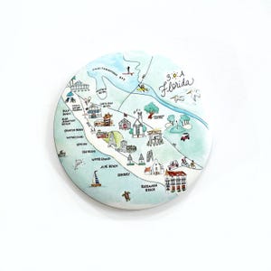 May include: A round, illustrated map of the 30A Florida area, featuring watercolor-style drawings of landmarks and beaches. The map includes text labels for locations like Santa Rosa Beach and Rosemary Beach, with a light blue and white color scheme.