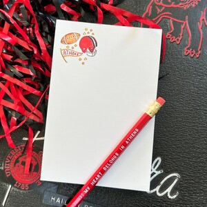 May include: A white notepad with a red and white football helmet, a football, and a red banner with the word "ATHENS" on it. The notepad has a red pencil with the text "MY HEART BELONGS IN ATHENS" on it.