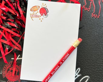 Könnte beinhalten: Ein weißer Notizblock mit einem rot-weißen Footballhelm, einem Football und einem roten Banner mit dem Wort "ATHENS" darauf. Der Notizblock hat einen roten Bleistift mit dem Text "MY HEART BELONGS IN ATHENS" darauf.