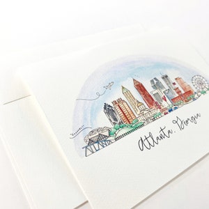 Pode incluir: Ilustra&ccedil;&atilde;o de aquarela do horizonte de Atlanta, Ge&oacute;rgia. A ilustra&ccedil;&atilde;o apresenta um arco-&iacute;ris sobre a cidade, com edif&iacute;cios, uma roda gigante e um avi&atilde;o voando por cima. O texto "Atlanta, Ge&oacute;rgia" est&aacute; escrito em it&aacute;lico na parte inferior da ilustra&ccedil;&atilde;o.
