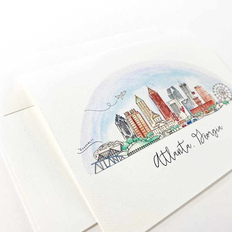 Pode incluir: Ilustra&ccedil;&atilde;o de aquarela do horizonte de Atlanta, Ge&oacute;rgia. A ilustra&ccedil;&atilde;o apresenta um arco-&iacute;ris sobre a cidade, com edif&iacute;cios, uma roda gigante e um avi&atilde;o voando por cima. O texto "Atlanta, Ge&oacute;rgia" est&aacute; escrito em it&aacute;lico na parte inferior da ilustra&ccedil;&atilde;o.