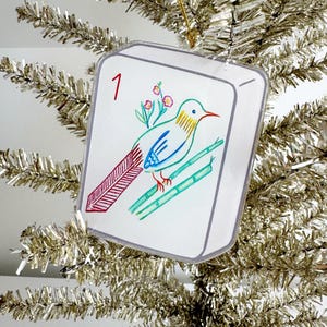 May include: A clear, square ornament with a hand-drawn bird design. The bird is perched on bamboo stalks, with a red tail and a blue body. The number "1" is in the upper left corner. The ornament hangs from a gold string on a silver tinsel tree.