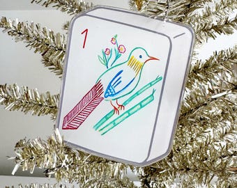 May include: A clear, square ornament with a hand-drawn bird design. The bird is perched on bamboo stalks, with a red tail and a blue body. The number "1" is in the upper left corner. The ornament hangs from a gold string on a silver tinsel tree.