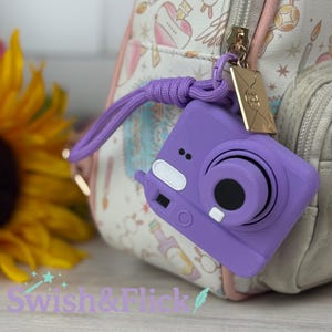 Puede incluir: Un charm de cámara morada con una correa de muñeca a juego está unido a una mochila. El charm tiene una etiqueta rectangular dorada. Un girasol y otras flores están en el fondo. El texto "Swish & Flick" está en la parte inferior.