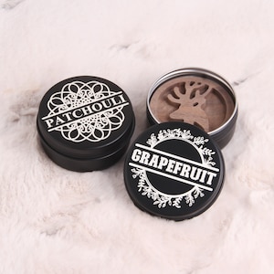 May include: Two round black metal containers with lids. One container has a white floral design and the text "GRAPEFRUIT". The other container has a white mandala design and the text "PATCHOULI".
