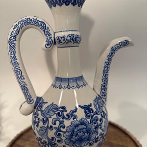 May include: White and blue ceramic pitcher with a lid. The pitcher features a curved handle and spout, adorned with blue floral and scrollwork patterns. The base has a large blue floral design.