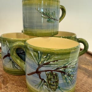 May include: A set of four ceramic mugs with a nature-inspired design. Each mug features a hand-painted pine branch with a pinecone against a blue and green background. The mugs have a yellow interior and green handle.