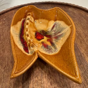 May include: A vintage ashtray with a unique, organic shape. The ceramic ashtray features a warm, earthy color palette with shades of brown, yellow, red, and white. The design includes a central well and a series of cigarette rests.
