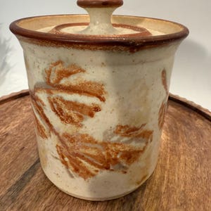 May include: A ceramic jar with a lid, featuring a beige base with brown leaf patterns. The rim and lid are accented with a darker brown. The jar is sitting on a wooden surface.
