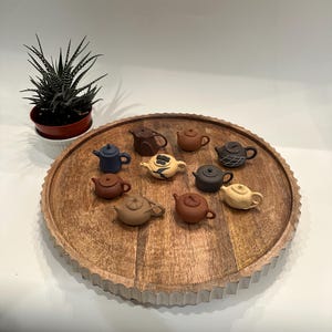 May include: A collection of ten small, colorful ceramic teapots arranged on a wooden tray with a scalloped edge. The teapots are in various shades of brown, blue, and cream.